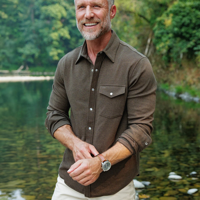 A smiling man with a gray beard stands outdoors by a calm lake, wearing a brown long-sleeve button-up shirt with a chest pocket and light-colored pants.