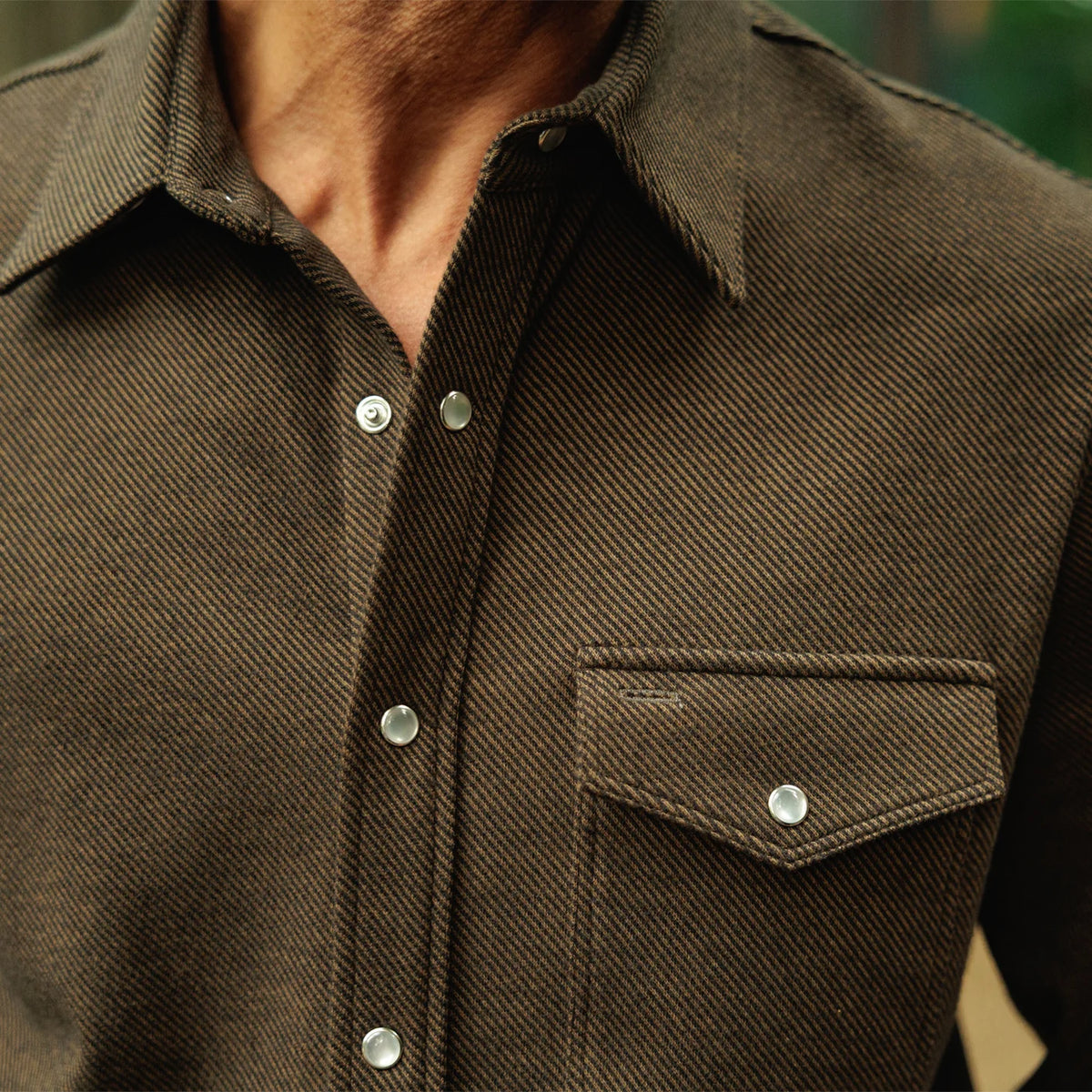 Close-up of a person wearing a dark brown textured button-up shirt with a chest pocket and silver snap buttons.