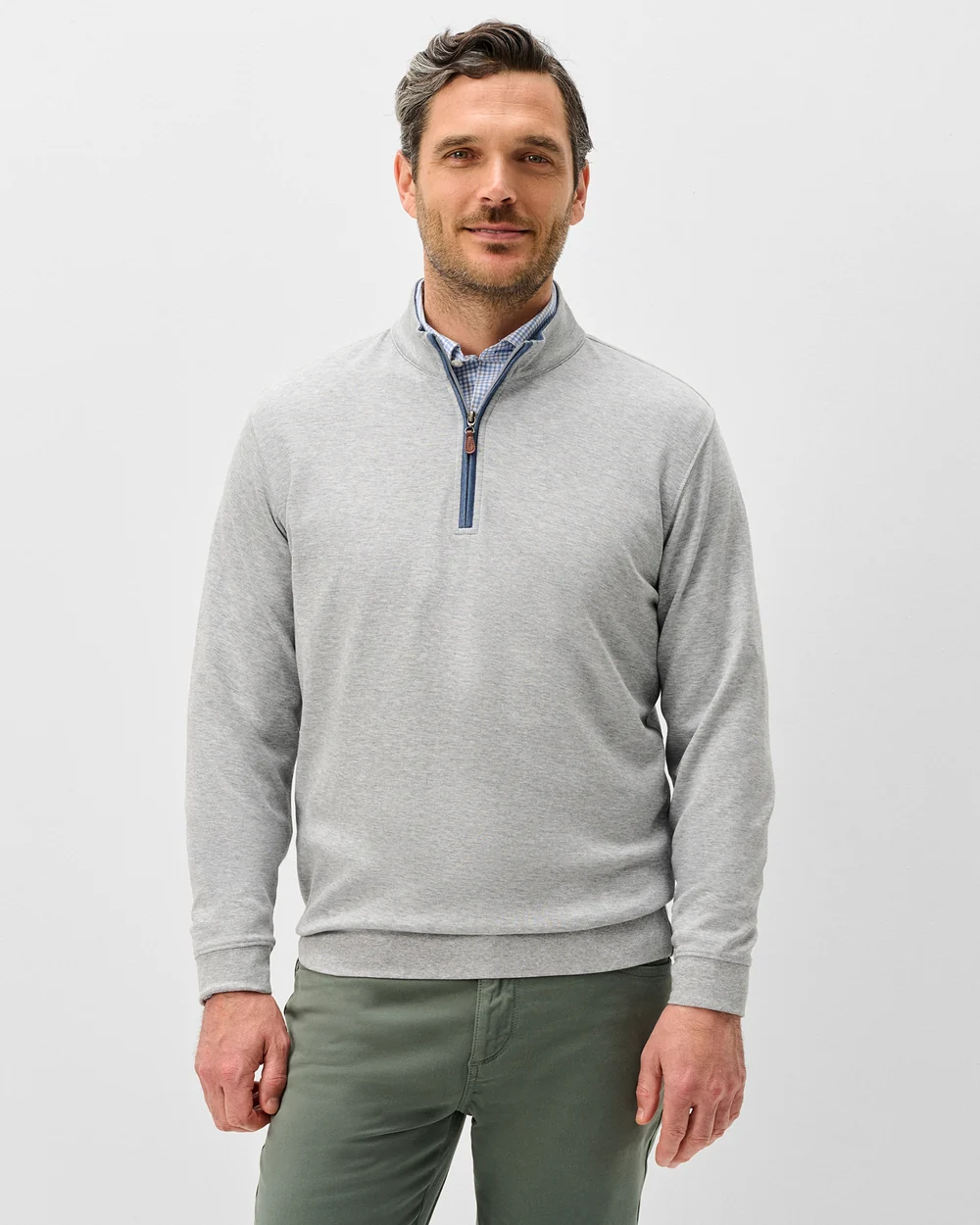 A man wearing a light gray quarter-zip pullover sweater over a blue patterned collared shirt and olive green trousers, standing against a light gray studio background.