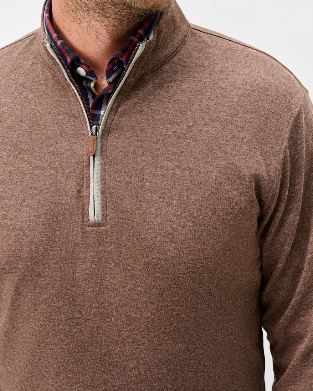 A close-up of a man wearing a brown heathered quarter-zip pullover sweater, layered over a red and blue plaid collared shirt. The sweater has a silver zipper and a small brown leather pull tab.