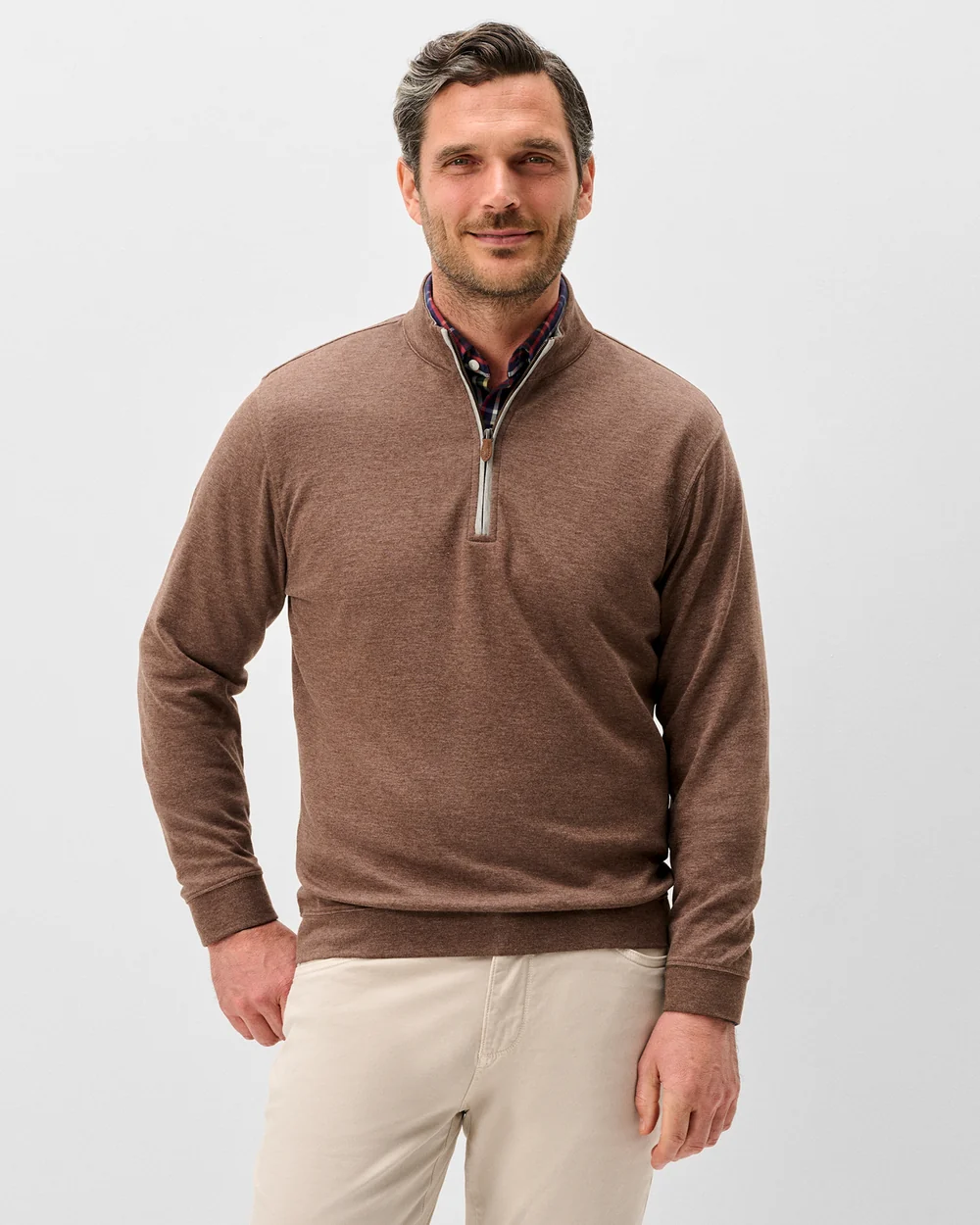 A man wearing a brown quarter-zip knit sweater over a patterned collared shirt and beige trousers, photographed against a light gray studio background.
