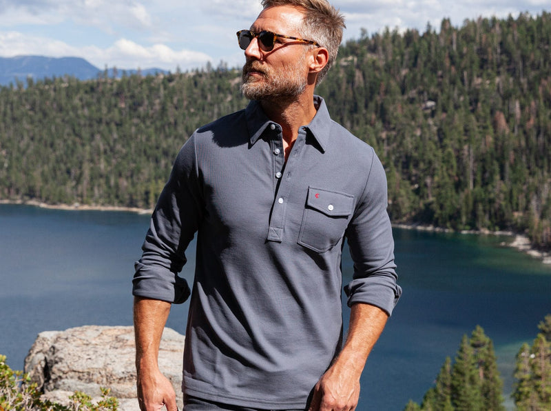 Man wearing sunglasses and a gray shirt standing in front of a scenic lake and forest.