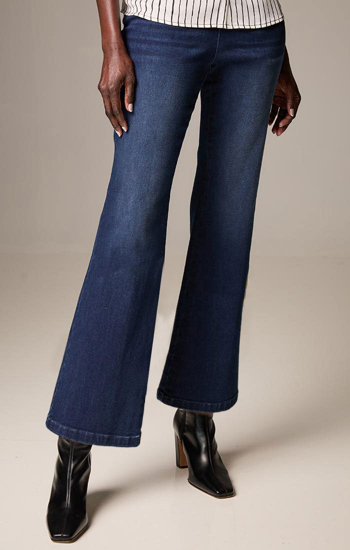 A person wearing dark blue flare jeans with a striped top and black ankle boots, shown from the waist down against a plain beige studio background.